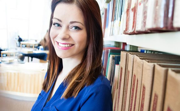 student looking at camera and smiling with back against bookshelf in library