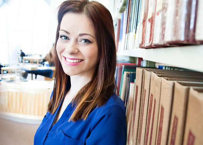 student looking at camera and smiling with back against bookshelf in library