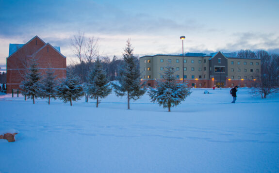 SSM Campus. Winter, evening. Snow covered residence building and person walking across campus through a path of snow lined with pine trees.